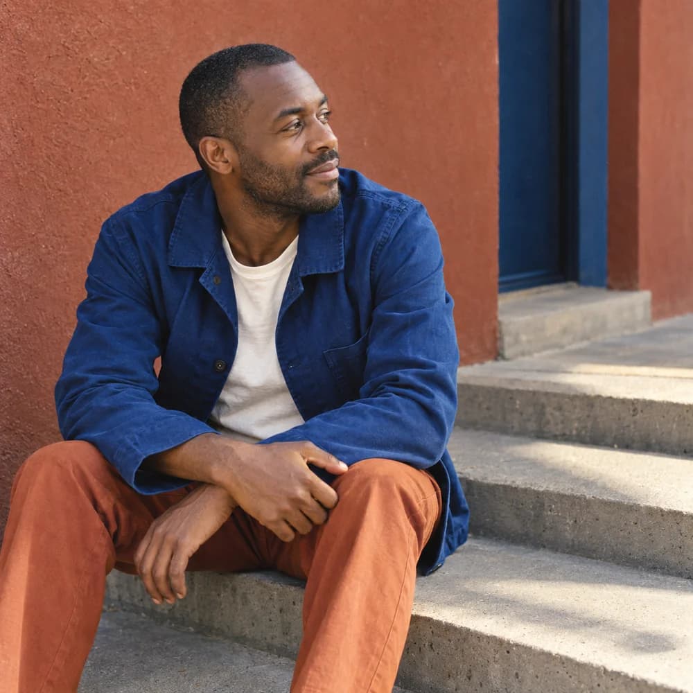 Illustrative portrait of a man in a cobalt-blue jacket sitting on steps outside a studio.
